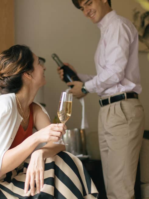 Person smiling and holding a glass of champagne while another person opens a bottle