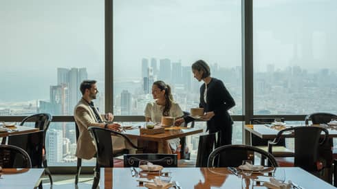 Several people meet in an Art Deco dining room overlooking the Bahrain cityscape. 