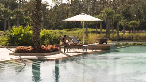 Outdoor swimming pool where woman lounges on patio chair under palm tree 
