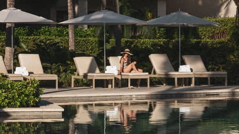 Person sitting on a poolside lounge chair under an umbrella at a tropical resort.