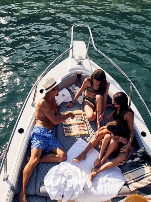 A family lounges in the bow of a luxurious boat amid towels and blankets, playing backgammon, the sun's glow on teal waters. 