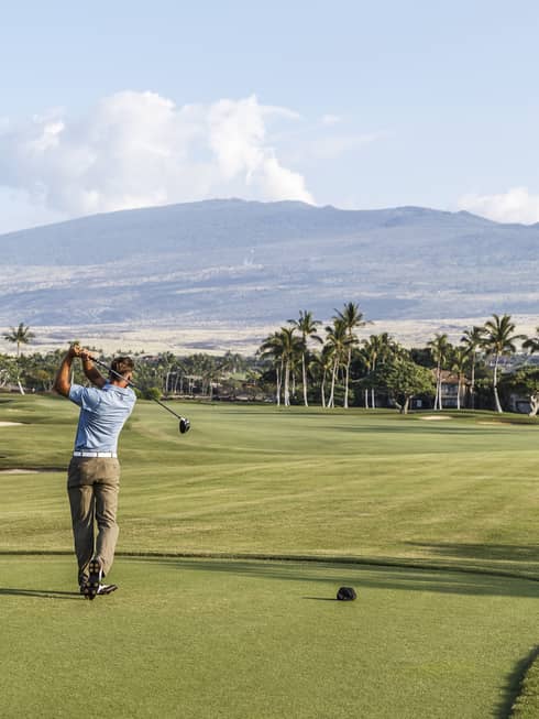 Back of man swinging golf club on course green with mountains in distance 