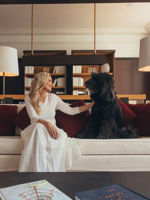 A person and a dog sit on a white couch with red pillows in a well-lit room with dark walls, a white ceiling and large windows