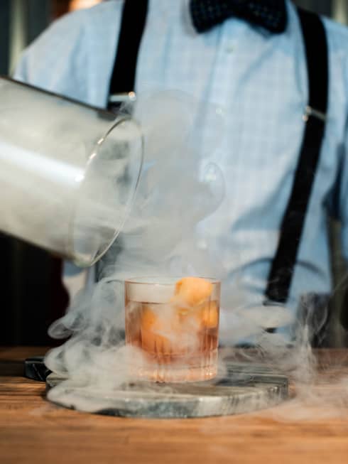 Smoke swirls around a cocktail as a bartender removes a lid 