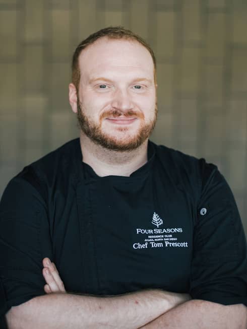 A male chef crossing his arms in black chef clothes in front of a brick wall.