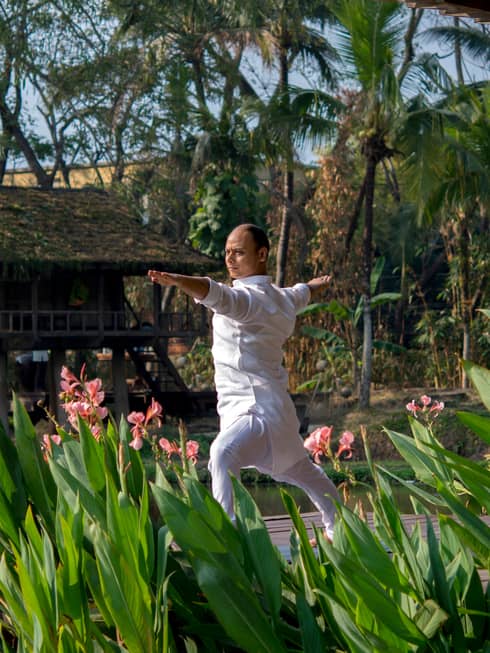 Resident yogi Dheera wearing white in yoga pose with arms outstretched by tall flowers, leaves