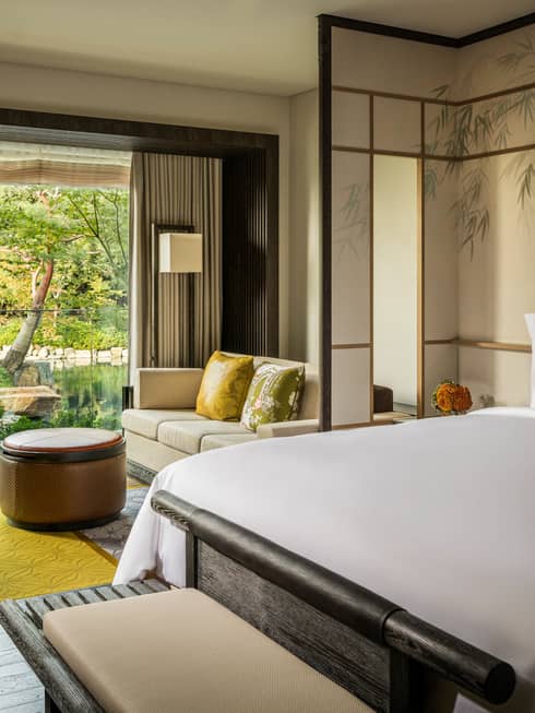 Guest room with a king bed, sofa with green cushions, round ottoman and large window overlooking a pond garden