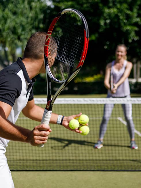 Couple playing tennis on court, man prepares to serve tennis balls to woman ready to swing
