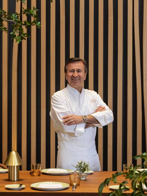 A chef in a white shirt and trousers stands in front of a pin-striped wall surrounded by foliage.