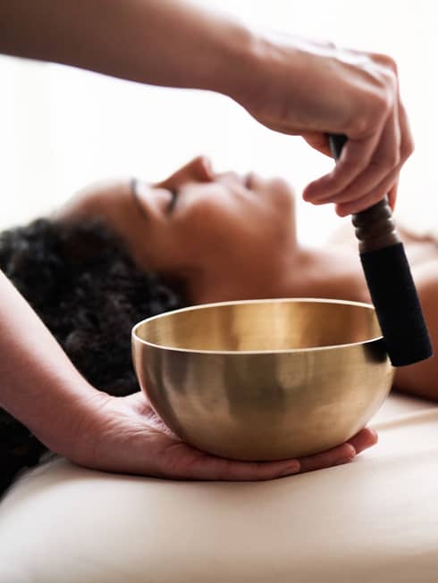Woman with eyes closed lies on massage table in as vibrational sound therapy is performed with a Tibetan singing bowl