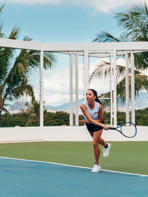 Front view of a player in follow-through after serving the ball on an outdoor tennis court, palm trees and mountains beyond.