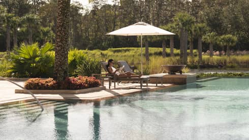 Outdoor swimming pool where woman lounges on patio chair under palm tree 
