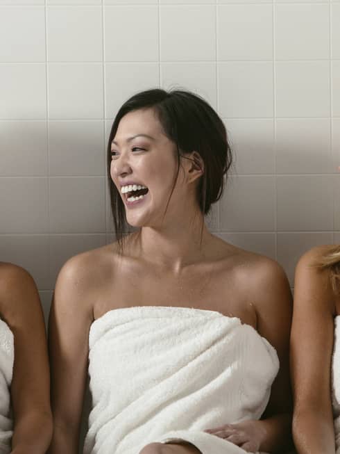 Three women wearing white towels sit side-by-side, laughing