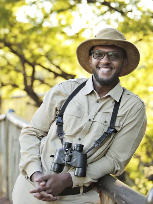 A man in beige clothes and a hat outside with binoculars.