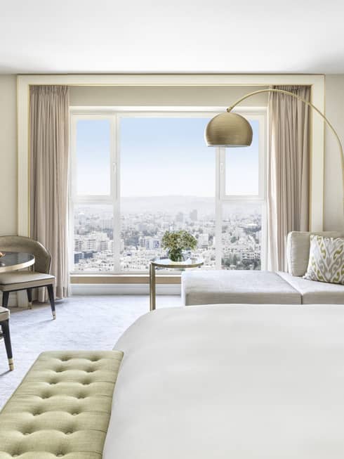 Hotel room with white king bed, light green cushioned bench, two taupe chairs, chaise, wall of windows