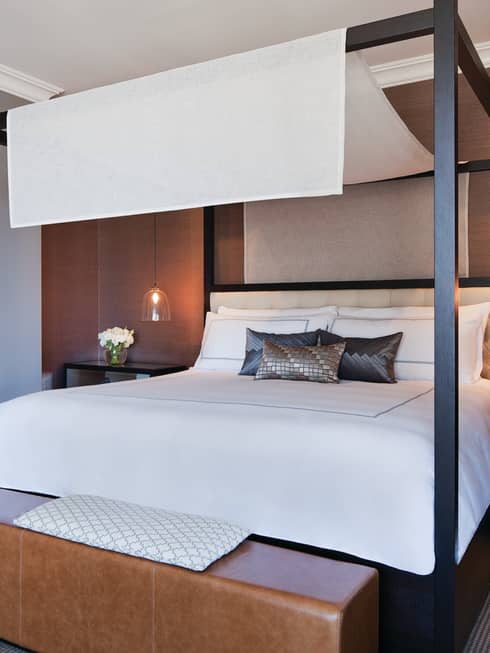 Poster bed with white linen canopy, silk accent pillows, brown leather bench at foot