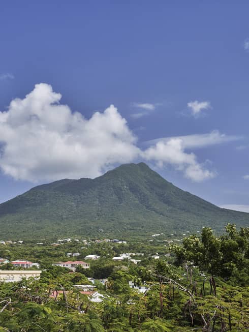 A large mountain in the background of a forested area with buildings poking out of the trees.