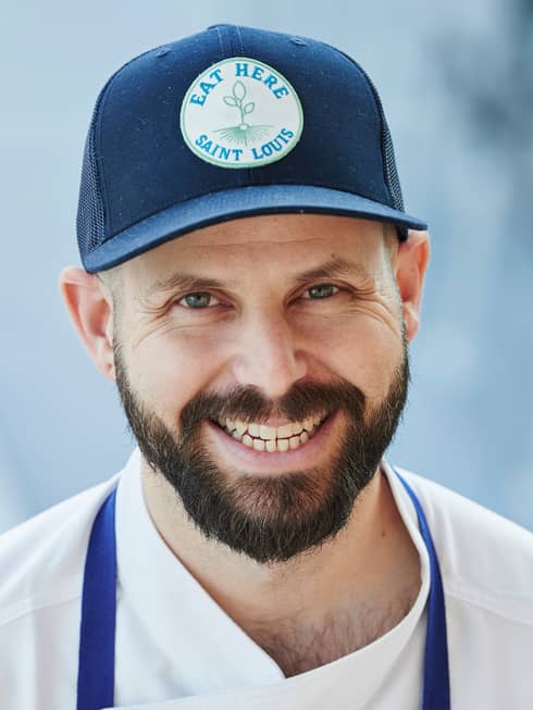 A male chef with a short bear in a baseball hat and blue apron smiling outside.