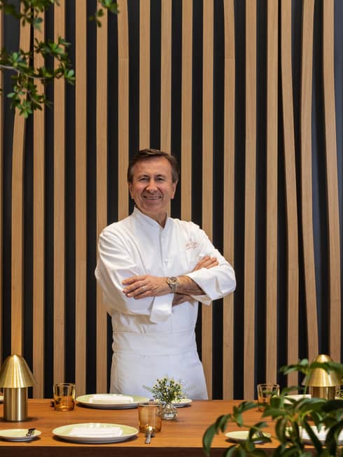 A chef in a white shirt and trousers stands in front of a pin-striped wall surrounded by foliage.
