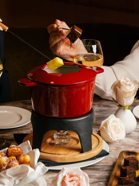 Two people sit at a table dipping fruit slices into a red pot of chocolate fondue