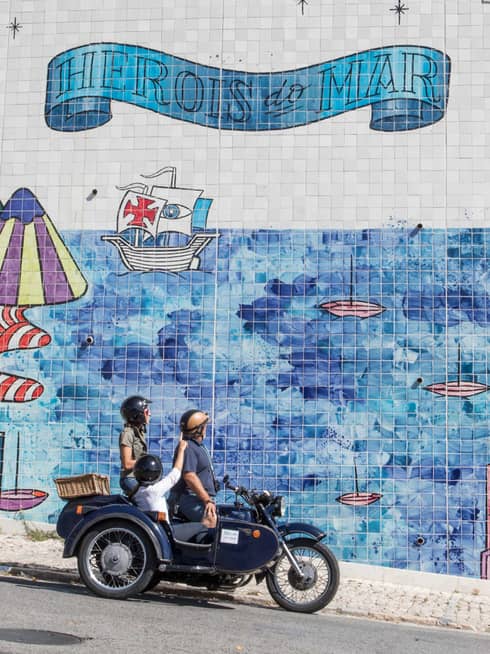 Two adults and a child stop their motorcycle to look at a city mural made of tiles