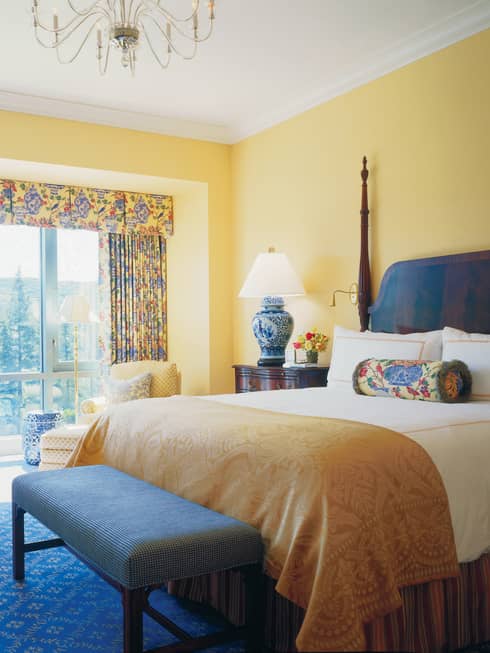 Deluxe Room hotel bed with gold blanket, bench, desk, windows with bright curtains