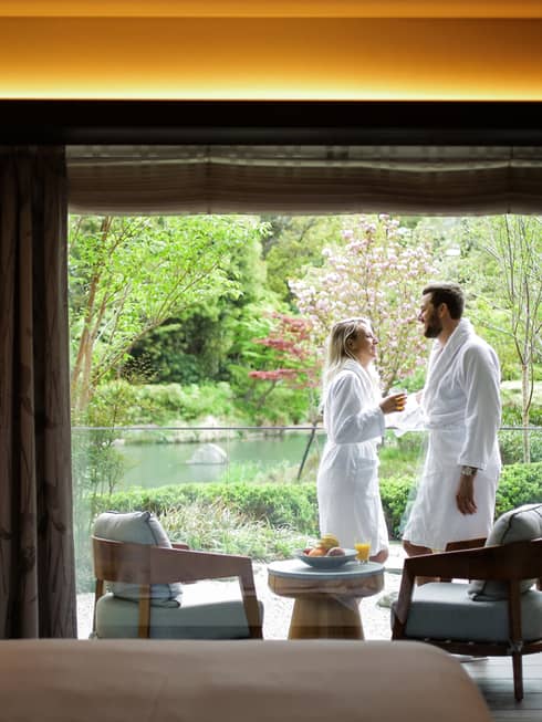 Smiling couple wearing white bathrobes on sunny balcony past hotel room bed