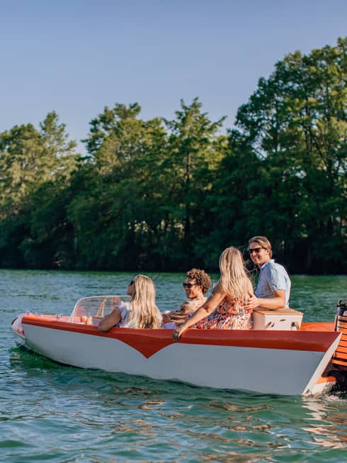 Two smiling couples with picnic baskets cruise through a river lined with trees in a red and white vintage boat.