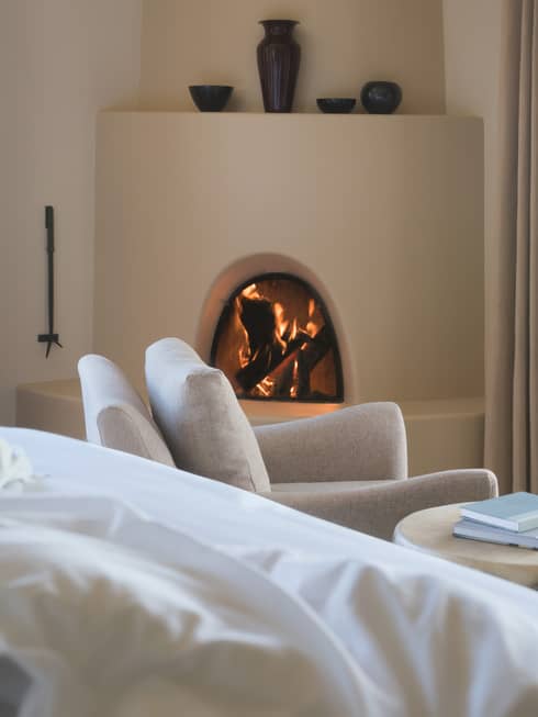 Hotel bed pillows, white armchair in front of curved clay fireplace, vases on mantle