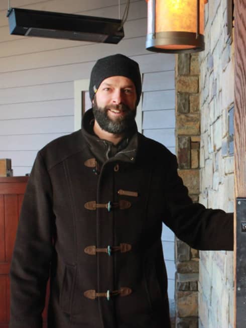 A man in a black and brown coat and beanie holding a wooden door with glass window open.