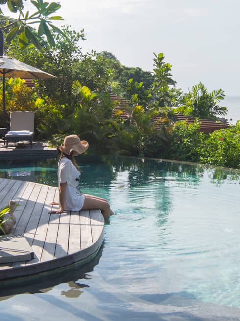 Woman wearing sunhat sits on villa patio, dips legs in infinity swimming pool 