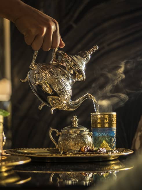 Hand pouring tea from Moroccan-style teapot into glass on tray