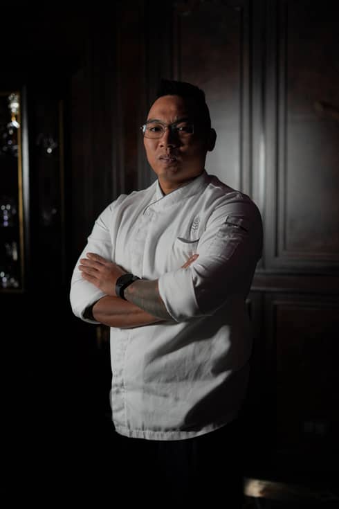 A chef wearing a white shirt stands with arms crossed in an interior space