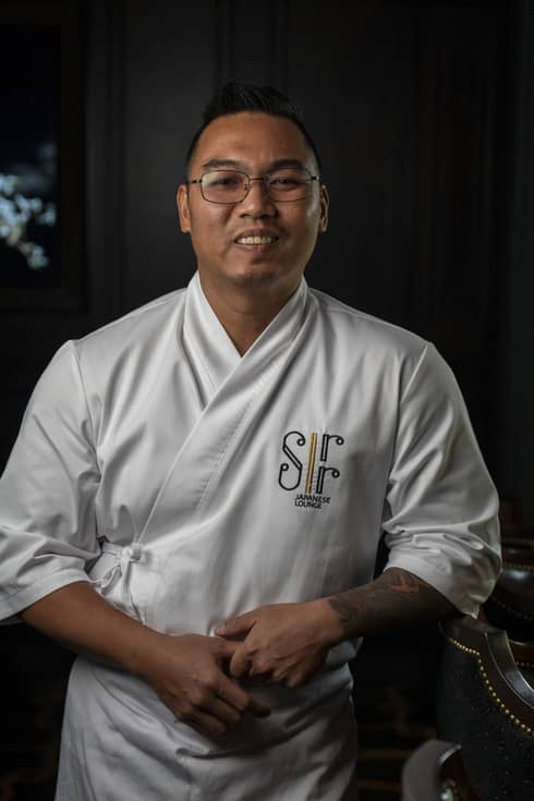 Chef with short dark hair and glasses, wearing a white chef's jacket and apron, stand smiling at the camera