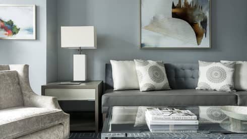 Midtown Suite long grey sofa with white accent pillows below abstract painting, table, lamp, armchair