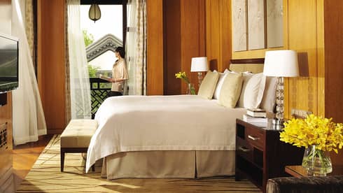 Woman stands in open window near hotel bed, bench, desk with bowl of oranges