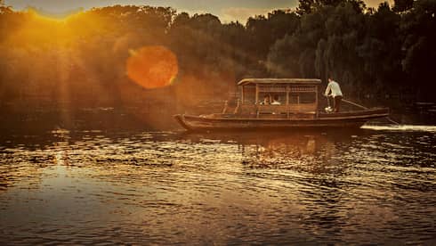 Man steers Chinese wooden rowboat on West Lake, into light of orange sunset