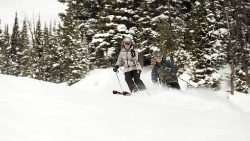 Man, woman ski down snowy hill under trees