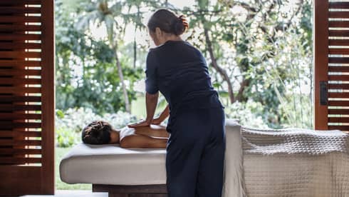 Spa staff massages woman's bare shoulders as she lies under sheet on massage table