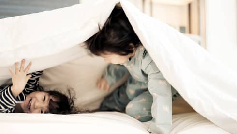 Two young children in pyjamas laugh, play under white blanket on bed