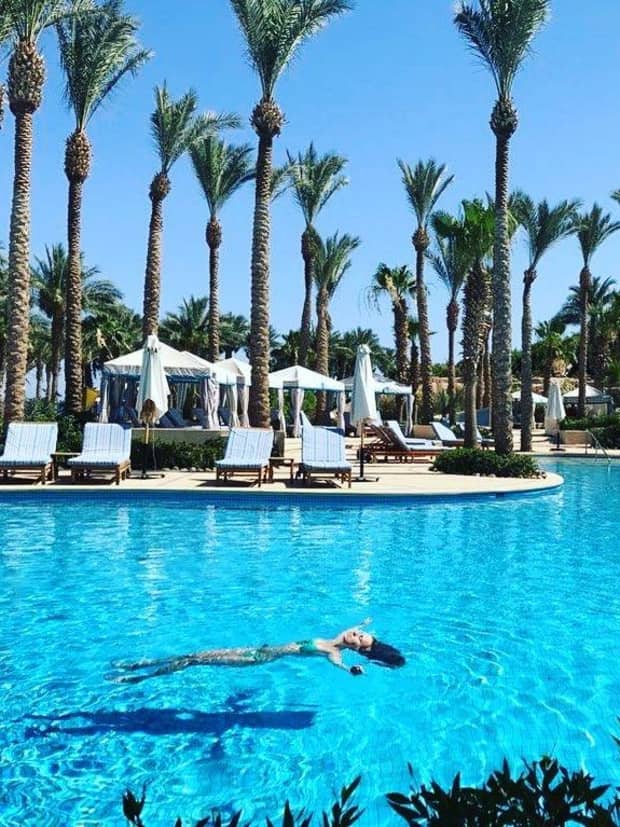 Woman floats on back in outdoor swimming pool, white cabanas, pool chairs and palm trees on deck