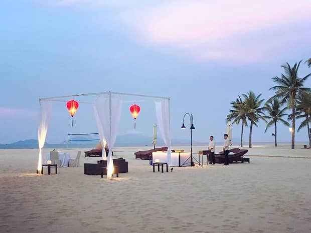 A romantic beach setup at dusk with red lanterns hanging from a white canopy, surrounded by lounge chairs, palm trees and soft lighting.