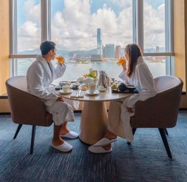 A couple dressed in white bath robes and slippers dine in front of a large window overlooking a city skyline