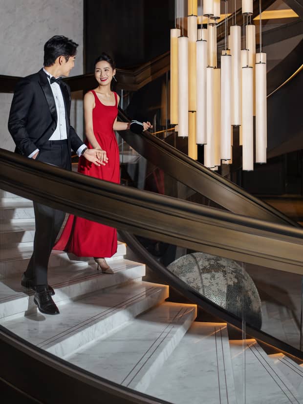 Man in a tux and woman in red gown walk down curved staircase, next to luxury lighting fixture, at Four Seasons Hotel Dalian