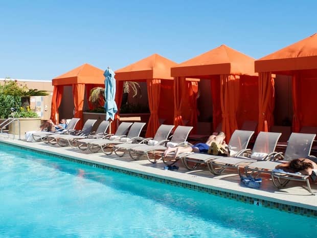 People suntan on lounge chairs lining outdoor swimming pool, bright orange cabanas