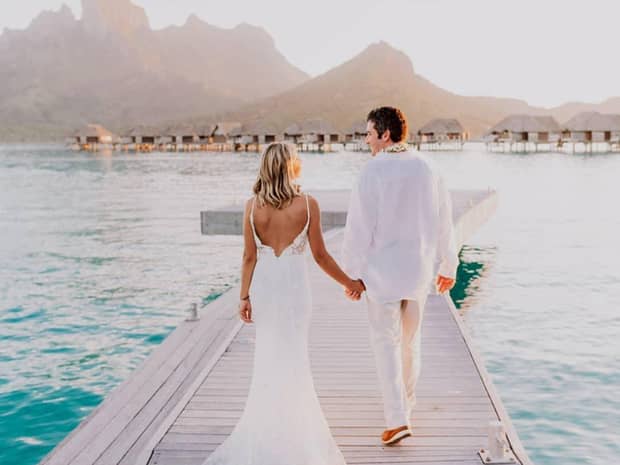 Back of bride and groom wearing white walking across platform over lagoon