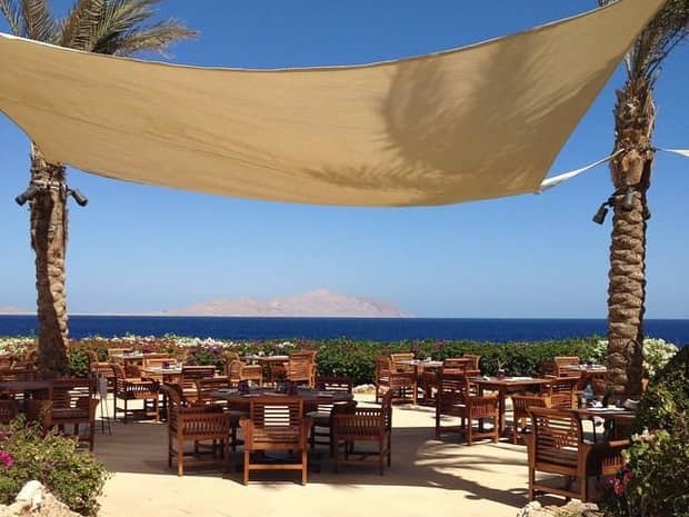 Large beige canopy connected to palm trees draped over outdoor dining tables, sea in background