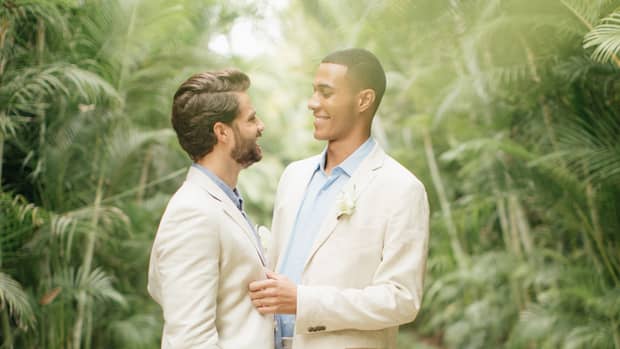 A gay couple is talking and embracing under a path of palm trees.