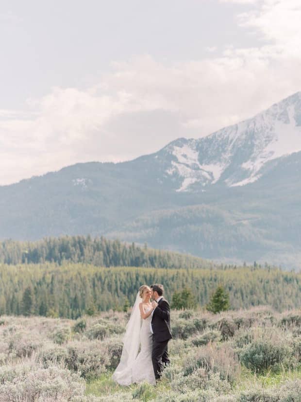 Couple dressed in wedding attire embracing in a scenic mountain field with lush greenery and snow-capped peaks in the background 