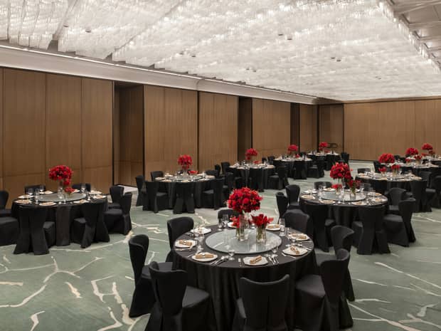 A spacious ballroom with wooden walls, with several black banquet tables set up with plates, glassware, linens and red floral arrangements